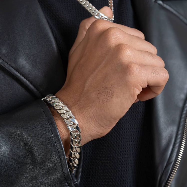 A close-up of a hand wearing the 12mm Handmade Miami Cuban Link Bracelet in 950 Silver, styled with a black leather jacket and dark shirt. This bold bracelet adds a striking, modern touch to their look.