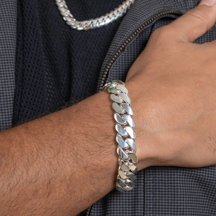 A close-up of a wrist wearing the 14mm Handmade Miami Cuban Link Bracelet in 950 Silver, featuring a small green detail. The person pairs it with a matching silver chain necklace and a dark textured jacket.