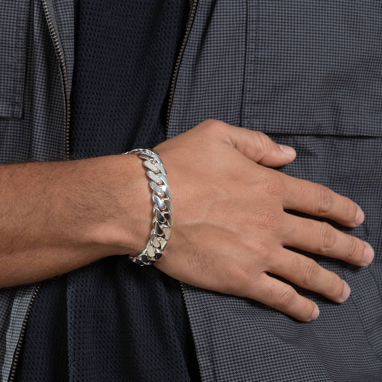 Someone wearing the 14mm Handmade Miami Cuban Link Bracelet - 950 Silver rests their hand on their chest over a gray checkered zippered jacket. The bracelet showcases large, flat, interlocking links and is expertly handmade.