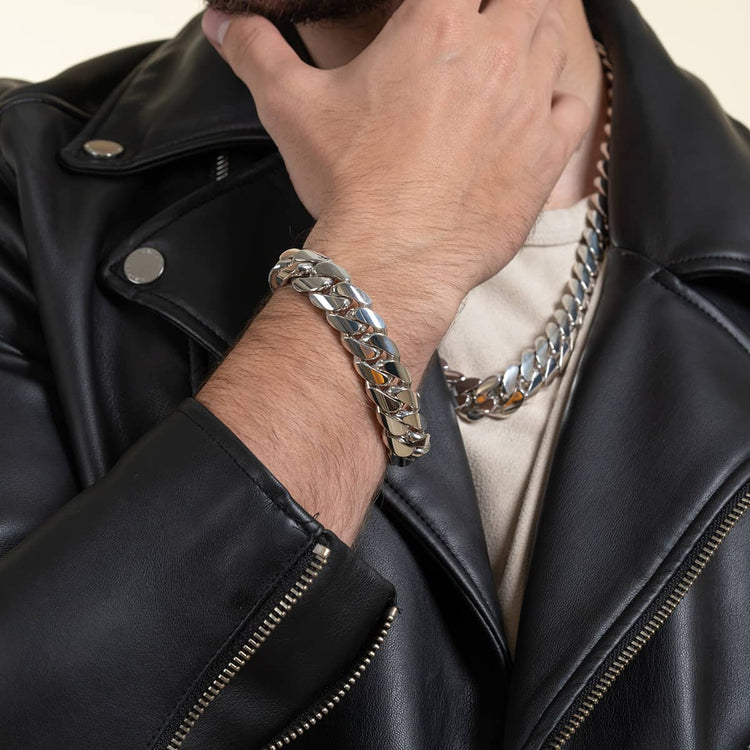 A person in a black leather jacket and cream shirt displays the 16mm Handmade Miami Cuban Link Bracelet - 950 Silver and a matching necklace, their hand thoughtfully touching their chin.