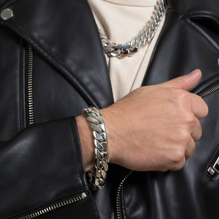 Wearing a black leather jacket and cream shirt, a person showcases the 16mm Handmade Miami Cuban Link Bracelet in 950 Silver on their wrist, paired with a matching silver chain necklace around their neck.