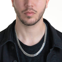 A man with short facial hair and a black shirt and jacket wears the Silver Cuban Link Chain - 10mm, shown from shoulders to mouth.