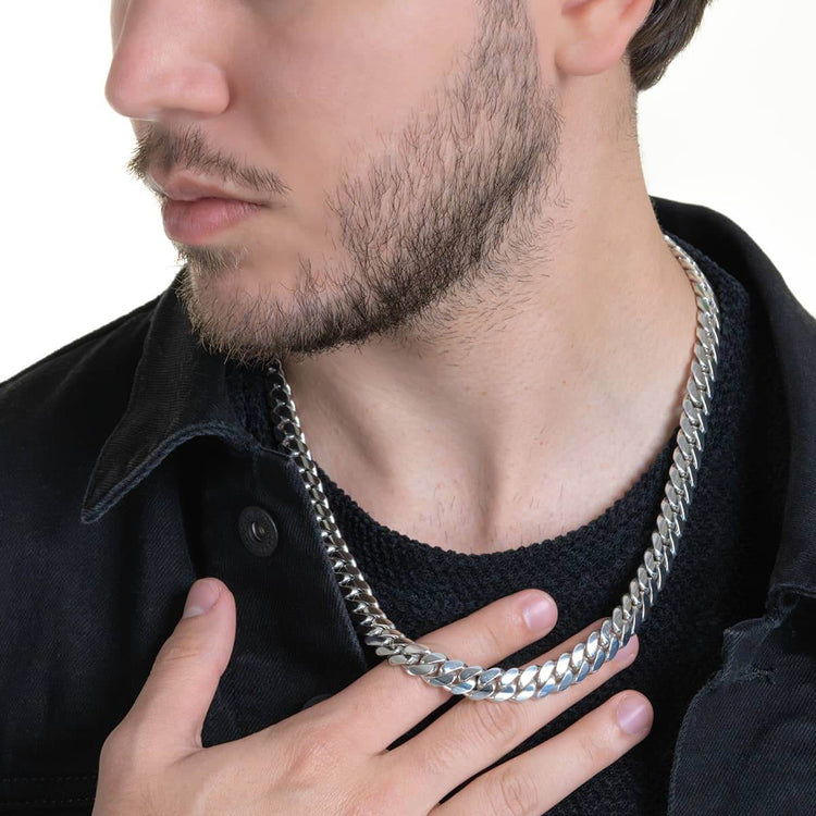 A close-up of a man with light facial hair wearing the 10mm Handmade Miami Cuban Link Chain in 950 silver, paired with a black textured shirt and jacket. He touches the necklace, highlighting its bold, premium craftsmanship.