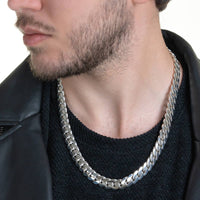 Close-up of a man wearing the Silver Cuban Link Chain - 12mm in 950 silver, styled with a black sweater and leather jacket; his face is partially visible with light facial hair.