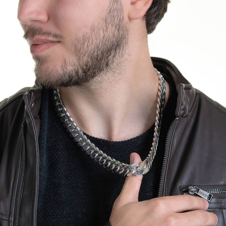 A man with short facial hair, wearing a black sweater and dark brown leather jacket, poses slightly turned while holding the 16mm Handmade Miami Cuban Link Chain - 950 Silver near his chest.