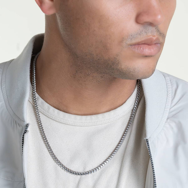 A close-up of a man wearing the Silver Cuban Link Chain - 5mm, styled with a cream t-shirt and light gray jacket. The shot focuses on his neck and chest, with his face partially visible.