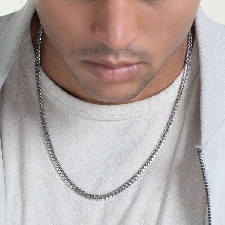 A person in a light shirt and jacket is shown from the shoulders up, wearing the Silver Cuban Link Chain - 5mm in 925 Sterling Silver around their neck.