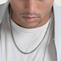 A person in a light shirt and jacket is shown from the shoulders up, with a close-up view of the Silver Cuban Link Chain - 5mm made of 925 Sterling Silver and plated with rhodium around their neck.