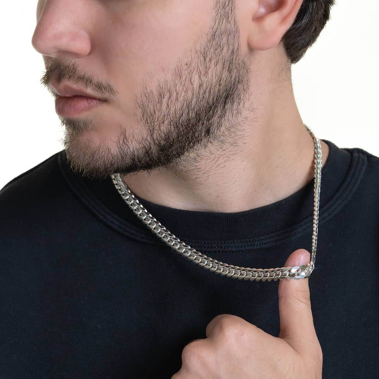 A man with a trimmed beard and mustache in a black shirt holds the 8mm Handmade Miami Cuban Link Chain - 950 Silver, pulling the silver chain slightly away from his neck against a plain, light background.