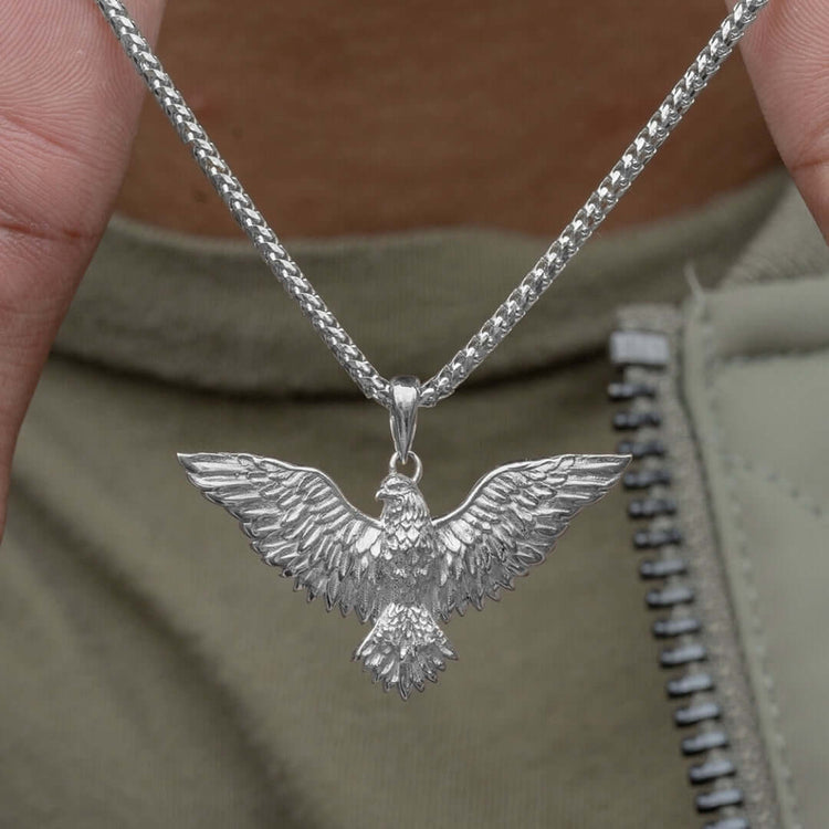 A close-up of the Silver Eagle Pendant, a silver chain necklace with a detailed eagle and outstretched wings, held in front of someone wearing an olive green shirt with a visible zipper.