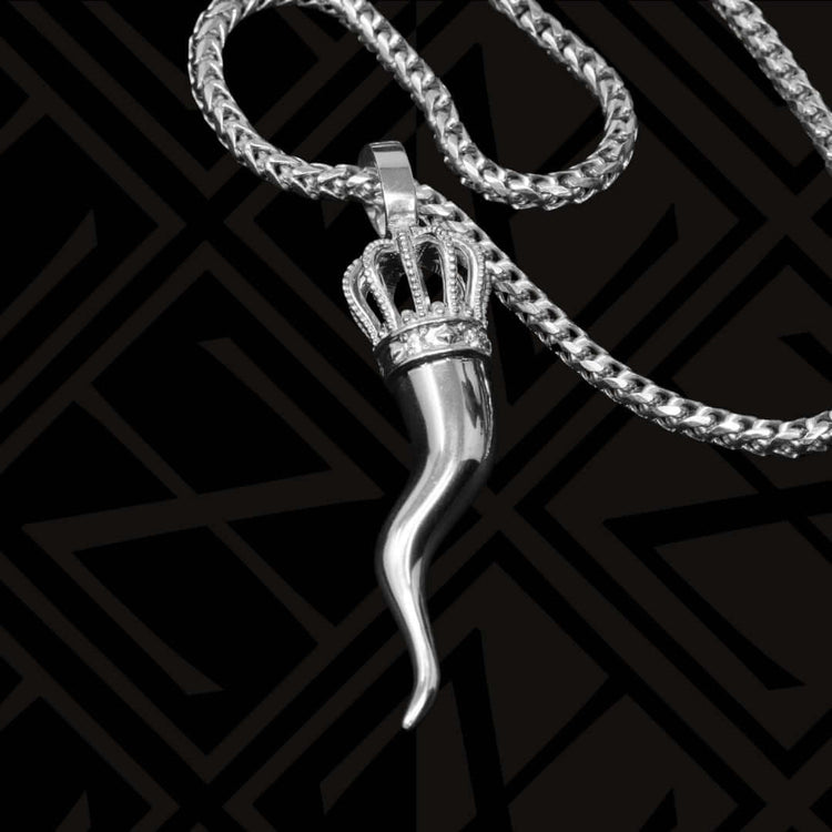 The Silver Italian Horn Pendant, adorned with a jeweled crown for added charm and protection, is elegantly showcased on a sleek black geometric patterned background.
