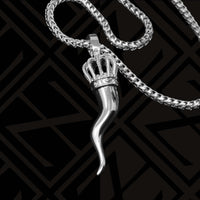 The Silver Italian Horn Pendant, adorned with a jeweled crown for added charm and protection, is elegantly showcased on a sleek black geometric patterned background.