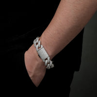 Someone wears the Silver Moissanite Cuban Link Bracelet – 20mm, sparkling with gems on their wrist as their hand rests in the pocket of black pants against a dark background.