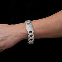 A person’s wrist adorned with the Silver Moissanite Cuban Link Bracelet – 20mm, featuring sparkling stones, set against a solid black background.