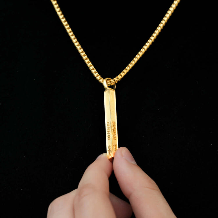 A hand holds the Solid Name Bar Pendant, a gold bar necklace with personalized engraving, hanging from a gold chain against a black background.