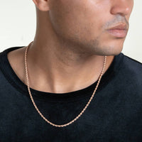 A man wears a black shirt and a Rope Chain - 2mm-5mm Solid Rose Gold necklace, shown from the chest up with his face partially visible, looking to the side.