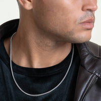 A man wearing the Franco Chain - 1.8mm-6.5mm Solid White Gold, a black shirt, and a brown leather jacket is shown from the shoulders up with his face partially visible and looking to the side.