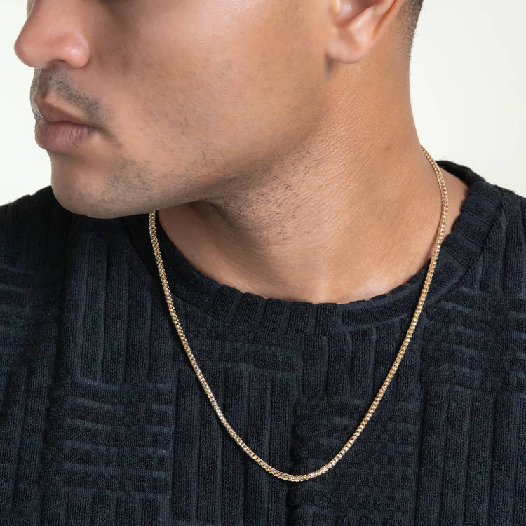 A person wears a textured black shirt and the Franco Chain Two Tone Prism Cut - 3mm 10k Solid Gold necklace. The image, cropped to show the lower face and upper chest, highlights the elegant design of the chain.