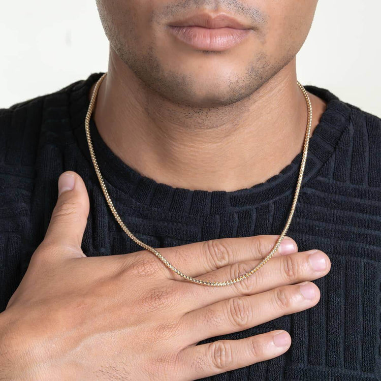 A person in a textured black shirt holds their hand to their chest, showcasing the shimmer of a 3mm two-tone prism cut Franco chain in 10k solid gold against light brown skin; only their lips and lower face are visible.