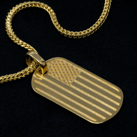 Show your patriotic spirit with the USA Flag Tag Pendant—a gold chain necklace featuring a rectangular engraved USA flag pendant displayed on a black fabric background.