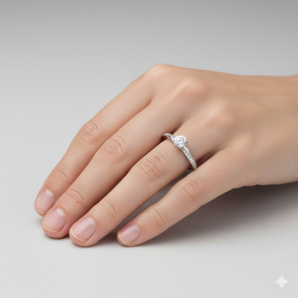 A hand with neatly manicured nails wears the Brilliance Eternity Lab Grown Diamond Engagement Ring, showcasing a round center stone and smaller diamonds along the band against a plain light gray background.