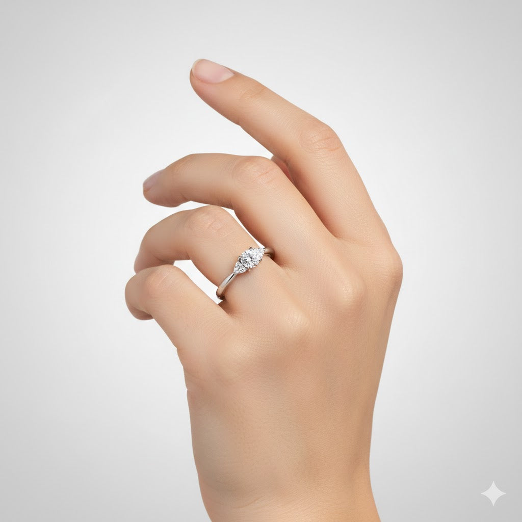 A hand with neatly manicured nails is shown against a light gray background, wearing the Round Brilliant Cut Diamond Engagement Ring on the ring finger.