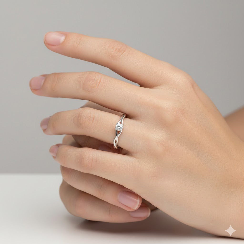 A hand with neatly manicured nails wears the Infinity Twist Lab Grown Diamond Solitaire Engagement Ring, featuring a single round lab-grown diamond in a twisted band. The background is plain and light gray.