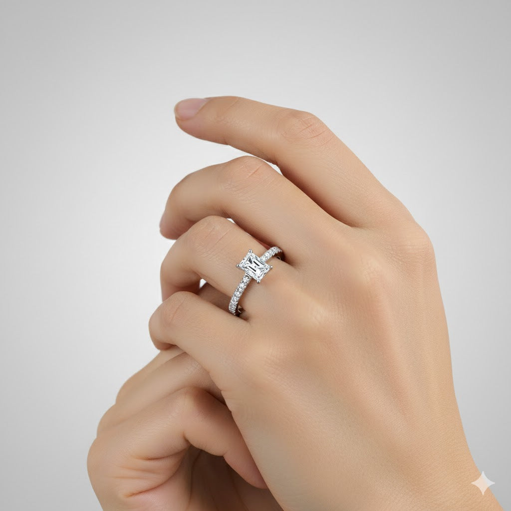 A close-up shows a hand wearing the Radiant-Cut Lab Grown Pavé Engagement Ring, featuring a square radiant-cut center stone and a pavé diamond band, set against a neutral background.