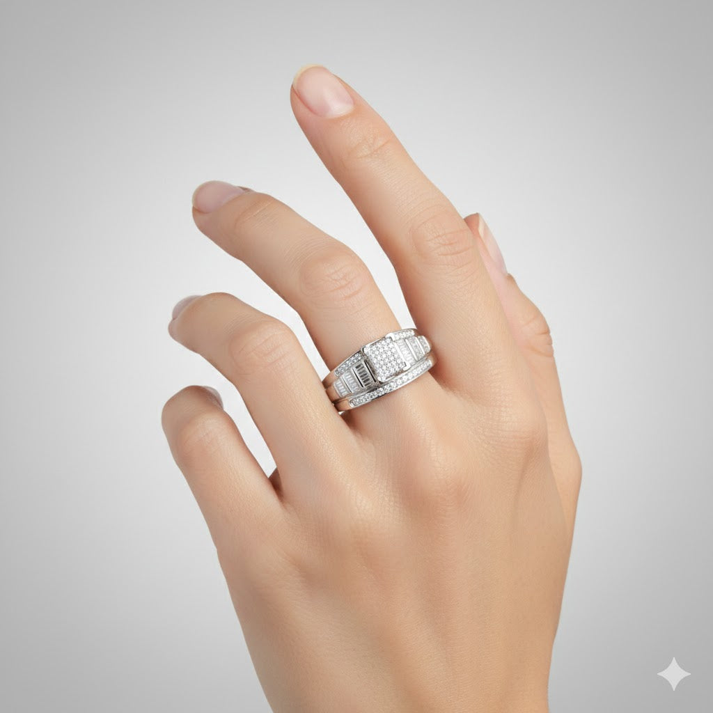 A hand with manicured nails wears the Sterling Silver Diamond Cluster Bridal Engagement Ring, featuring rectangular stones and a round diamond cluster, showcased against a plain light gray background.