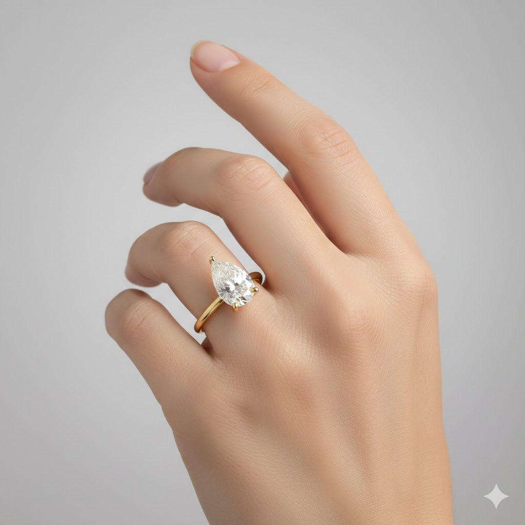 A hand with neatly groomed nails wears the 2.08ct Pear Lab Diamond Solitaire Engagement Ring in 14K yellow gold, featuring a stunning pear-cut lab-grown diamond against a plain light gray background.
