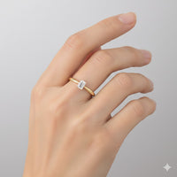 A hand with neatly manicured nails wears the 1/4CT Diamond Emerald Solitaire Ring 14K Gold on the ring finger, featuring an emerald-cut diamond, set against a plain light background.