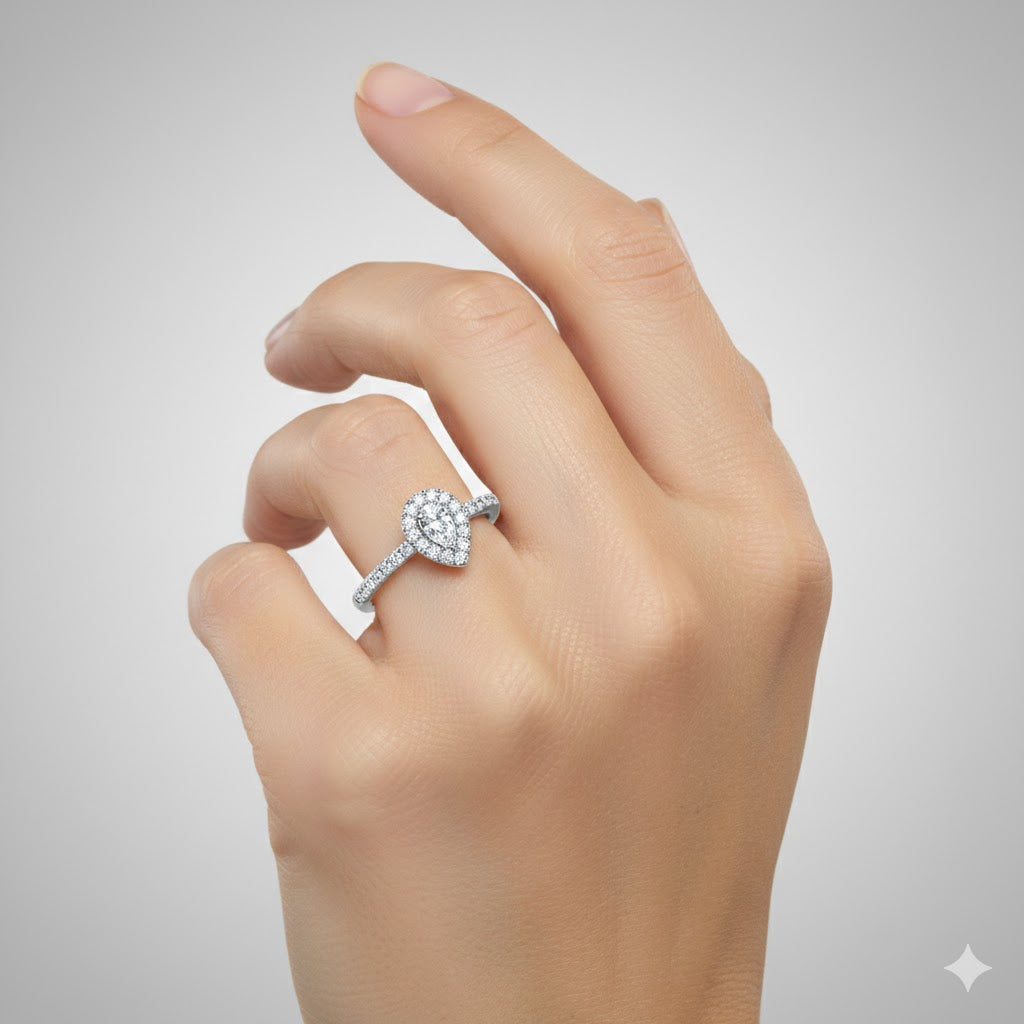 A hand with manicured nails is shown against a light gray background, wearing the Elegant Pear-Shaped Lab Diamond Halo Ring with a matching diamond band.