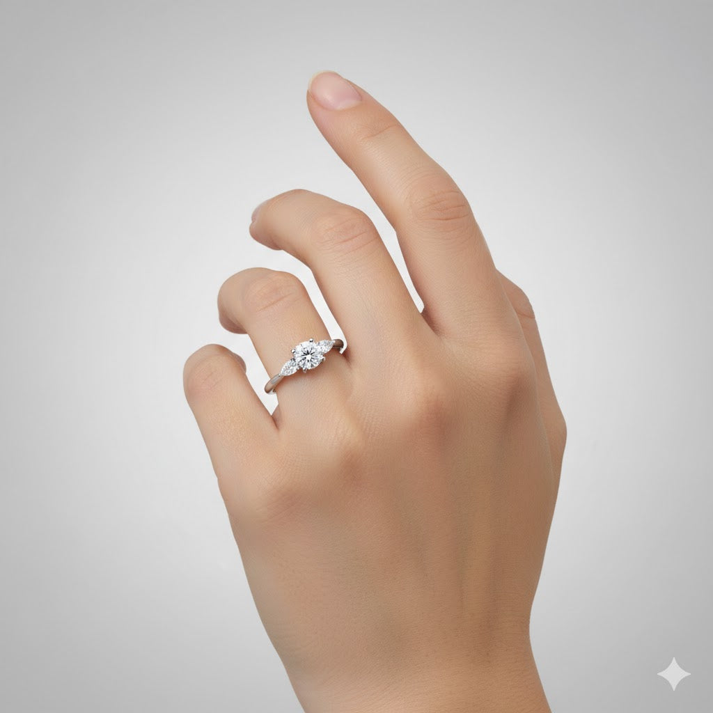 A hand with light skin is shown against a plain gray background, wearing the Three-Stone Round Lab Diamond Ring with Pear-Cut Side Stones on the ring finger.