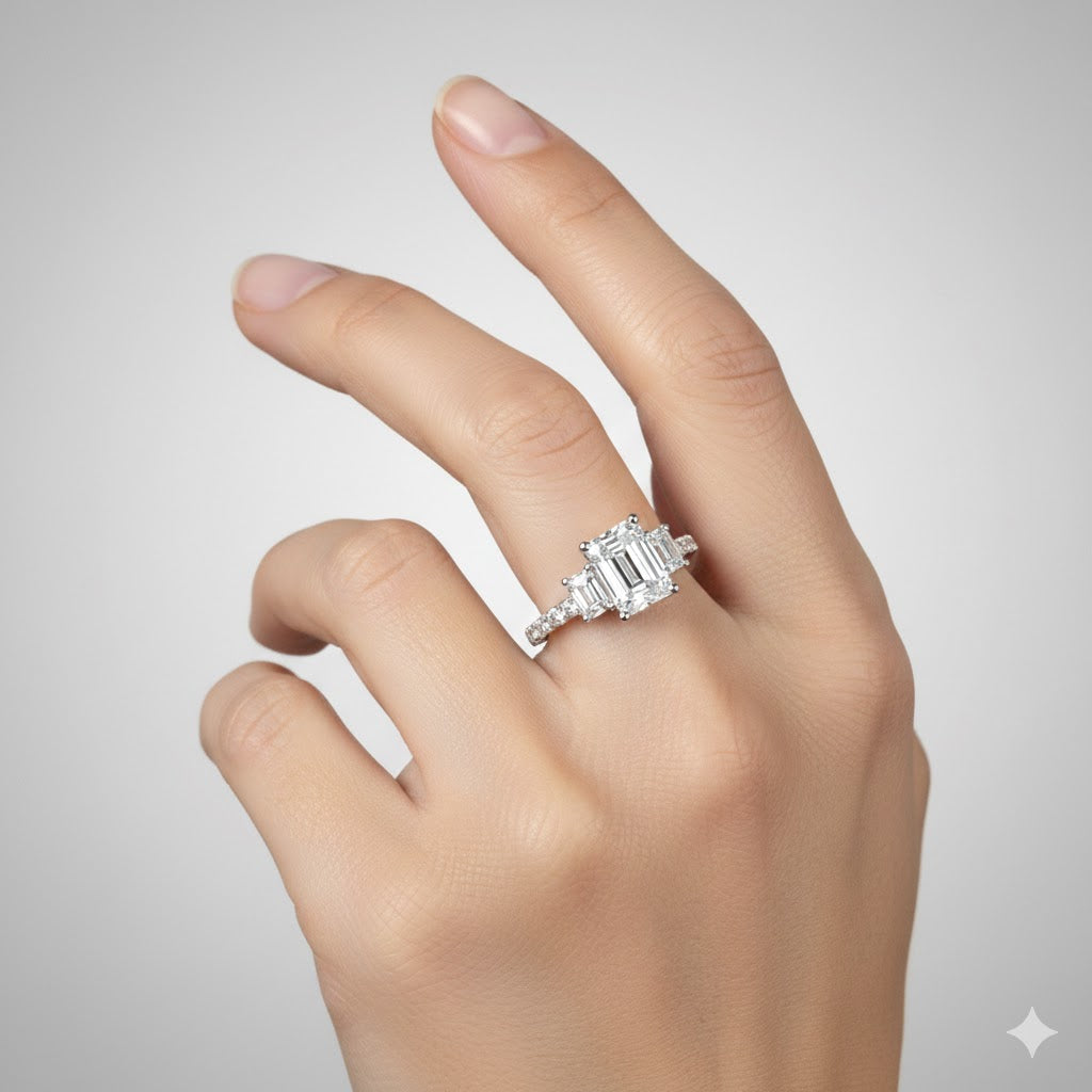 A hand with manicured nails showcases the 3.8 CTW 14k White Gold Three Stone Emerald Engagement Ring, featuring a large rectangular emerald flanked by two lab-grown diamonds, set against a simple light gray background.