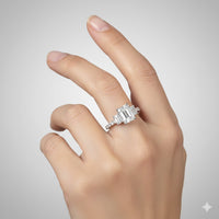 A hand with manicured nails showcases the 3.8 CTW 14k White Gold Three Stone Emerald Engagement Ring, featuring a large rectangular emerald flanked by two lab-grown diamonds, set against a simple light gray background.