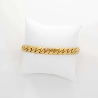 The Cuban Link Bracelet - 10mm Solid Gold is showcased on a white cushioned pillow, standing out against a clean white background with its thick, shiny design.