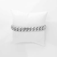 A thick, silver chain bracelet on a white cushion contrasts perfectly with the bold Cuban Link Bracelet - 11mm Solid Gold, showcasing its standout design.