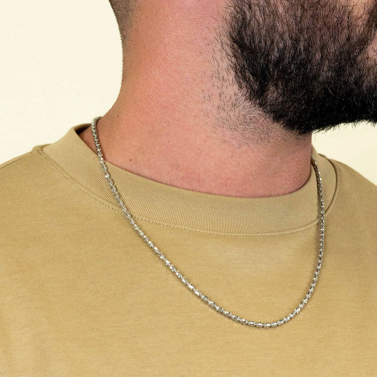 A close-up shows a person in a beige t-shirt wearing the 3mm Barrel Crystal Chain in 925 silver. Only the lower part of their bearded face is visible; their full face is out of frame.