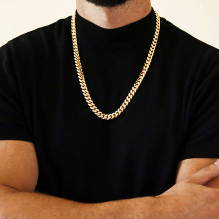 A person with crossed arms wears a black short-sleeve shirt and a bold men’s jewelry piece—the Cuban Link Chain 10mm in 14k Gold Bonded. Their face is not shown.