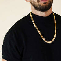 A man in a black t-shirt wears the Cuban Link Chain 10mm - 14k Gold Bonded, shown from the shoulders up against a plain light background—perfect for those seeking bold men’s jewelry.