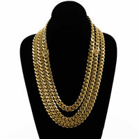 The Cuban Link Chain 12mm - 14k Gold Bonded is displayed on a black velvet bust with thick, layered gold chains featuring flat, interlocking links. Expertly crafted for lasting shine, against a plain white background.