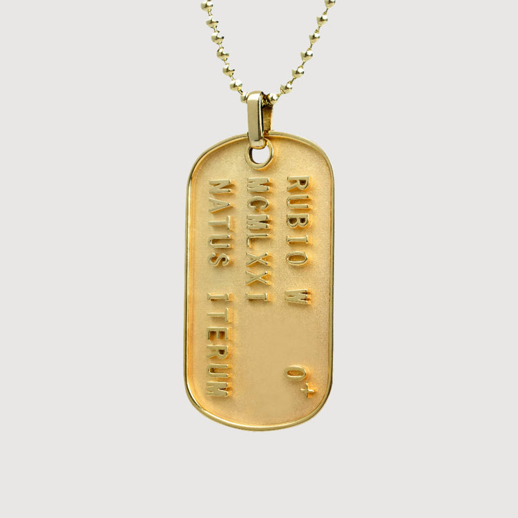 The Custom Military/Dog Tag Pendant features raised lettering: “RUBIO M,” “MCMLXXI,” “NATUS ITB ERIUM,” and “O+” on a gold dog tag with a ball chain, displayed against a plain white background.