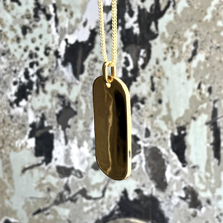 The Custom Military/Dog Tag Pendant features a smooth, rounded rectangular gold pendant hanging from a fine gold chain, displayed against a mottled black, white, and gray background.