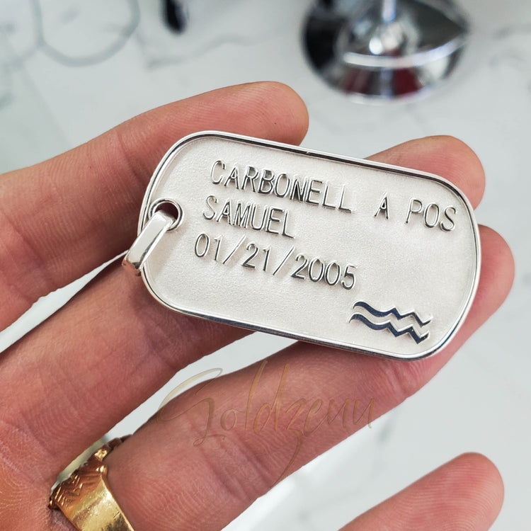 A hand holds a Custom Military/Dog Tag Pendant engraved with "CARBONELL A POS, SAMUEL, 01/21/2005" and a wave symbol. The blurred background with metallic objects inspires this personalized gold dog tag pendant.