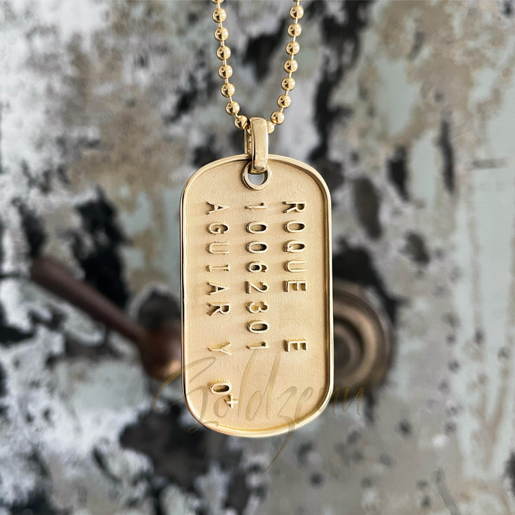 A Custom Military/Dog Tag Pendant engraved with "ROQUE 1006301 AGUIAR" hangs before a textured, weathered surface with peeling paint and a blurred brown object in the background.