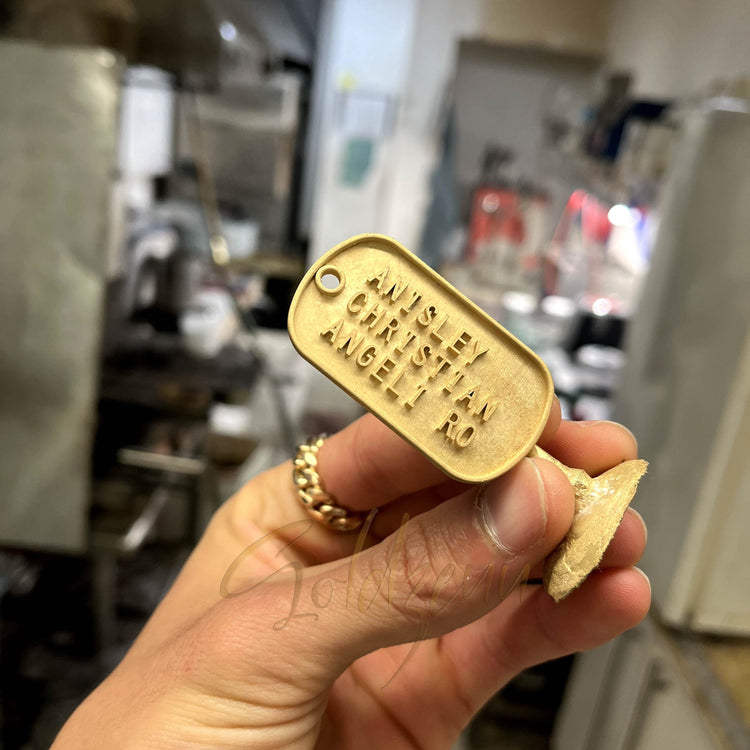 A hand holds the Custom Military/Dog Tag Pendant engraved with ANISEY CHRISTIAN AUGET 10 in a workshop, with tools and equipment blurred in the background, showcasing the pendant’s personalized craftsmanship.