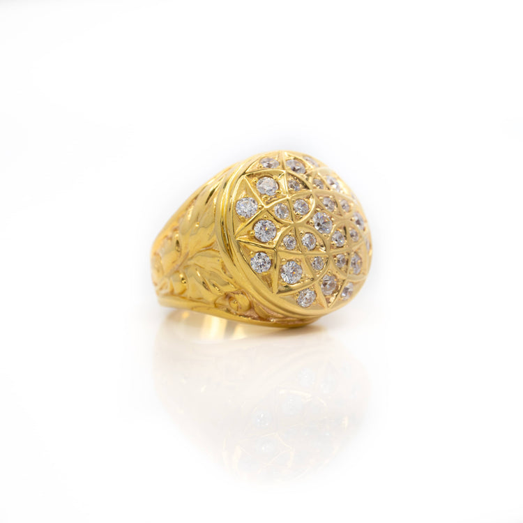 The Round Cluster Ring in Solid Gold features an intricately engraved band and a large round top adorned with multiple sparkling stones, shown on a white reflective surface.