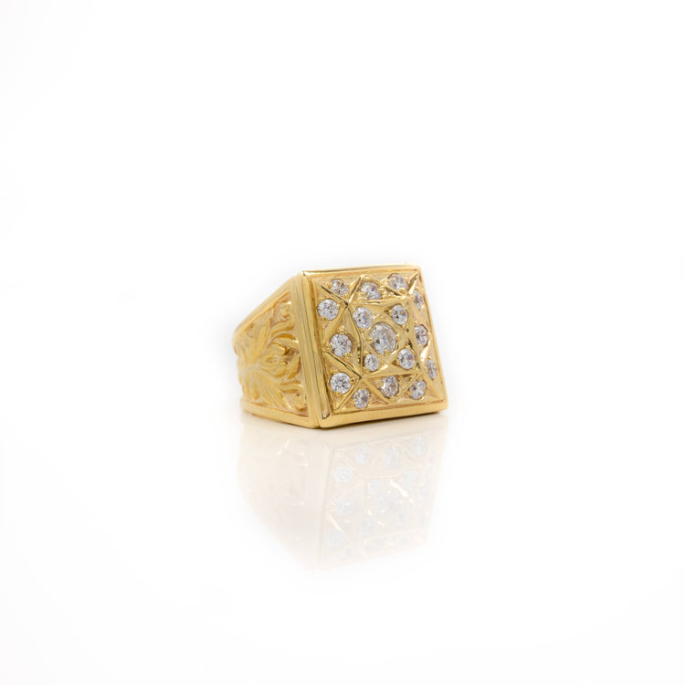 The Square Cluster Ring in Solid Gold features intricate engravings and a top set with multiple clear round gemstones, displayed on a white reflective surface—a stunning solid gold statement piece.