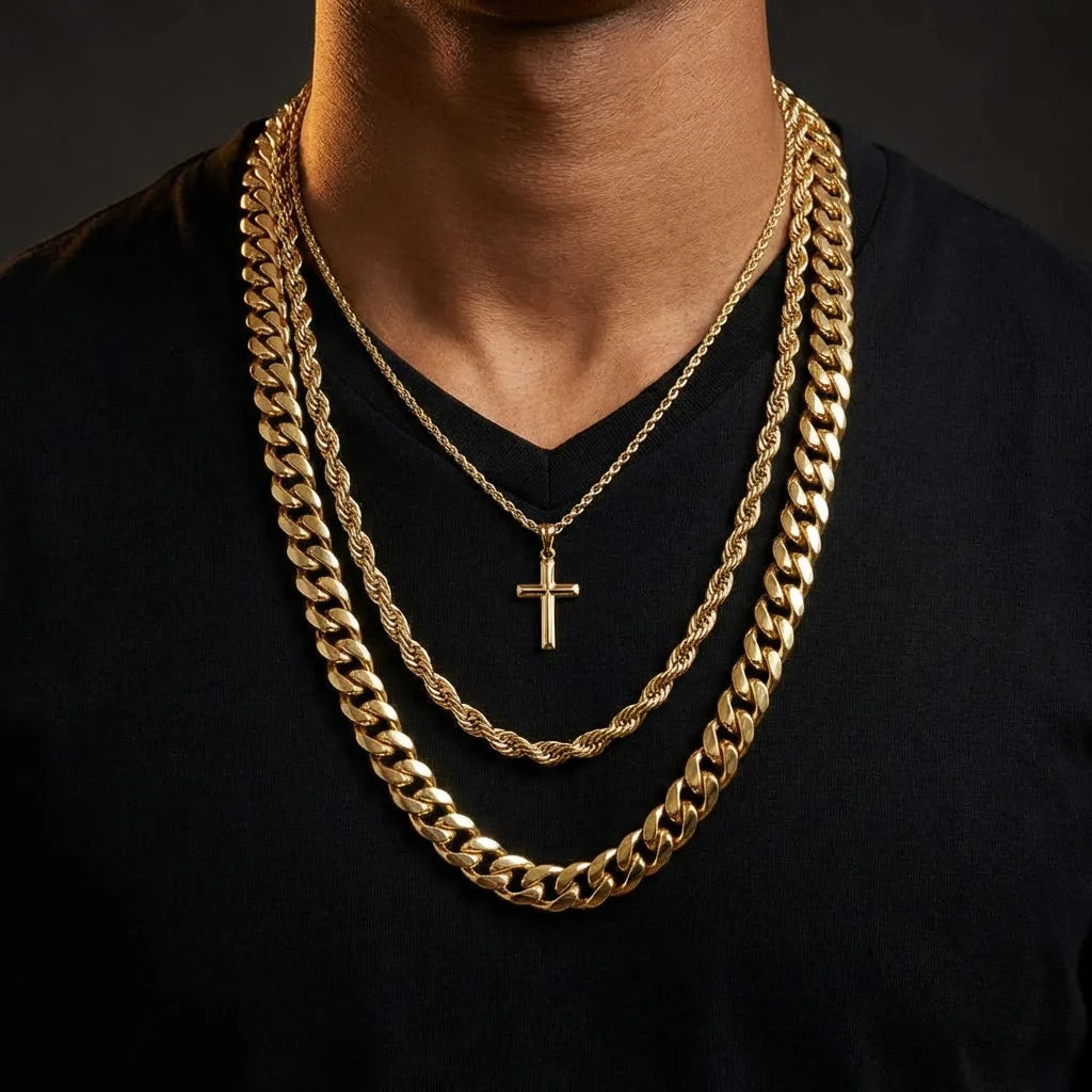 Man from neck to mid-chest wearing three layered gold chains: a thick 12mm Cuban link at 22 inches, a 6mm rope chain at 20 inches, and a thin 3mm chain with cross pendant at 18 inches, against a dark background with warm dramatic lighting