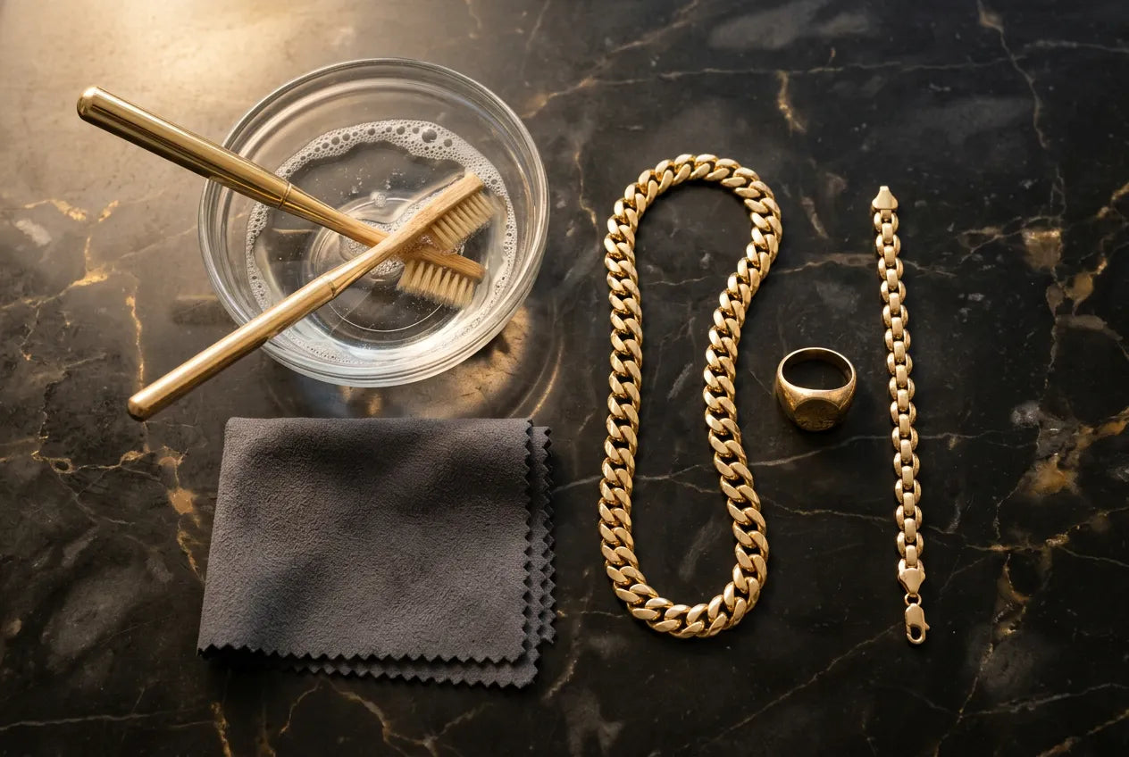 Gold jewelry cleaning supplies arranged on dark marble including warm water bowl, soft brush, and polishing cloth with gold Cuban link chain, ring, and bracelet